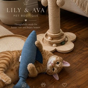 Peut inclure: Un chat tigré orange allongé sur un parquet en bois, tenant un jouet en forme de poisson en tissu bleu. Derrière le chat se trouve un griffoir en sisal beige avec une base en bois en forme de fleur et des petites billes. Texte LILY & AVA PET BOUTIQUE.