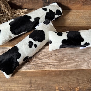 May include: Three black and white cow print fabric lumbar support pillows. The pillows are long and narrow and are made of a soft, plush fabric.