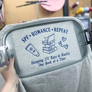 Op de afbeelding: Een lichtgrijze stof in een grijze borduurring met donkerblauw geborduurde tekst die SPF • ROMANCE • REPEAT en Escaping UV Rays & Reality One Book at a Time leest, met iconen van een zonnebrandcrèmetube, een stapel boeken en twee hartjes.
