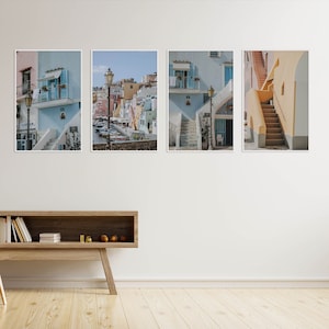 May include: Four framed art prints depicting colorful buildings and streets. The prints showcase a coastal town with blue, pink, and yellow buildings, stairs, and street lamps. The artwork is displayed above a wooden console with a leather chair.