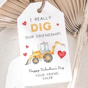 May include: White tag with the text "I REALLY DIG OUR FRIENDSHIP!" in gold and black. A yellow excavator with a heart-shaped bucket is featured. Red hearts are scattered around the text. The tag reads "Happy Valentine's Day, YOUR FRIEND, CALEB."