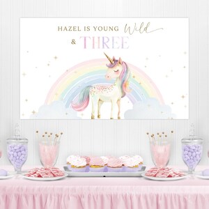 May include: A party table with a pink tablecloth, treats, and a banner that reads "Hazel is Young, Wild & Three" with a unicorn and rainbow design. The table includes cupcakes, cookies, and candy.
