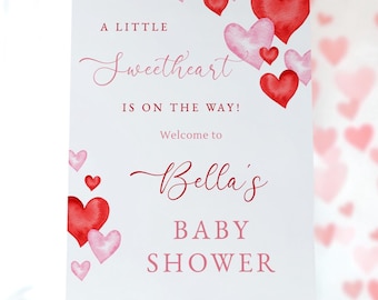 Könnte beinhalten: Ein weißes Schild auf einer Holzstaffelei mit dem Text "A Little Sweetheart Is On The Way!" und "Welcome to Bella's BABY SHOWER" in roter Schrift. Aquarellherzen in Rot und Rosa verzieren das Schild.