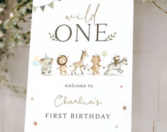 May include: A white sign on a wooden easel with the text "wild ONE" in gold script. Below, it reads "welcome to Charlie's FIRST BIRTHDAY" with illustrated animals. The sign has a green and gold color scheme.