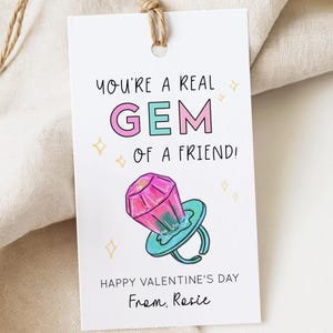 May include: A white gift tag with the text "YOU'RE A REAL GEM OF A FRIEND!" in black and pink. A cartoon ring with a pink gem and a teal band is featured. The tag also says "HAPPY VALENTINE'S DAY From. Rosie".