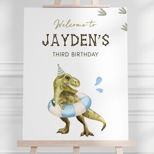 Puede incluir: Un letrero blanco sobre un caballete con el texto "Welcome to JAYDEN'S THIRD BIRTHDAY" en letras doradas y marrones. Una ilustración en acuarela de un dinosaurio verde con un gorro de fiesta y un flotador a rayas azules y blancas.