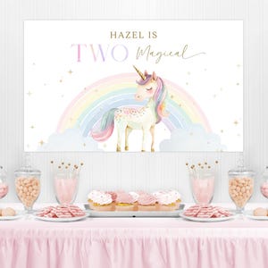 May include: A party table with a pink tablecloth is set with treats and decorations. A large banner with a unicorn, rainbow, and the text "Hazel is Two Magical" hangs in the background. Glass jars and plates hold candies, cookies, and cupcakes.