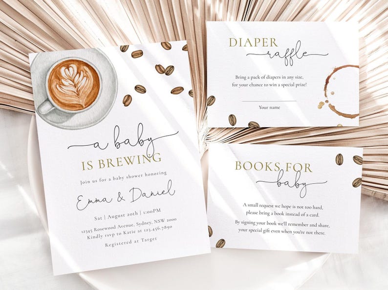 May include: A white card with gold lettering announcing a baby shower for Emma & Daniel. The theme is "a baby is brewing" and features a watercolor illustration of a cup of coffee. The card includes the date, time, and location of the shower. There are two additional cards for guests to fill out, one for a diaper raffle and one for a book request.