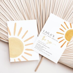 Peut inclure: Une carte d'invitation blanche avec un graphique de soleil jaune et le texte "LAILA'S FIRST TRIP AROUND THE SUN" et des d&eacute;tails pour une f&ecirc;te de premier anniversaire.