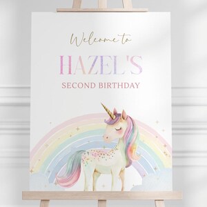 Puede incluir: Un letrero blanco sobre un caballete de madera con el texto "Welcome to HAZEL'S SECOND BIRTHDAY" en colores dorados y pastel. Un unicornio con melena y cola de arcoíris está frente a un arcoíris.