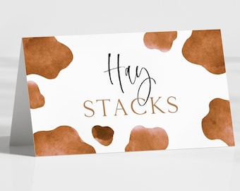 May include: A folded white card with brown cow spots and the words "Hay Stacks" in a script font. The card is angled, suggesting it is a place card or a small sign.