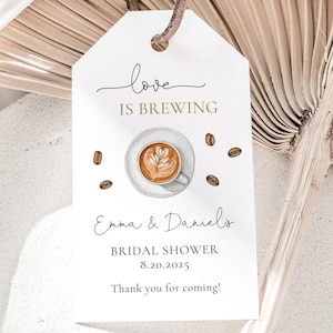 May include: White tag with the text "Love is Brewing" in black script font and gold lettering. A watercolor illustration of a cup of coffee with latte art is in the center. The text "Emma & Daniel's Bridal Shower 8.20.2025 Thank you for coming!" is below the illustration.