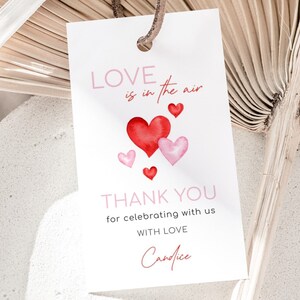 May include: A white gift tag with the text "LOVE is in the air" and "THANK YOU for celebrating with us WITH LOVE Candice". The tag features watercolor hearts in shades of red and pink, with a brown string.