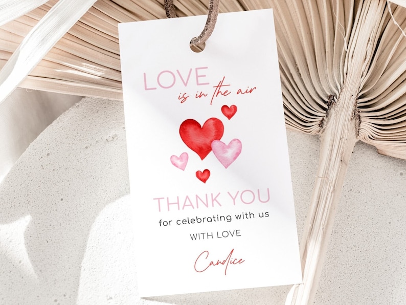 May include: A white gift tag with the text "LOVE is in the air" and "THANK YOU for celebrating with us WITH LOVE Candice". The tag features watercolor hearts in shades of red and pink, with a brown string.