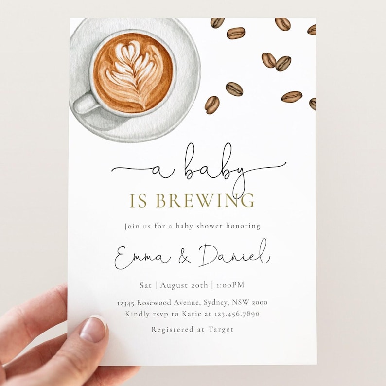 May include: A white card with a watercolor illustration of a cup of coffee with a latte art design. The text "a baby is brewing" is written in black cursive script with the word "brewing" in gold. The text "Join us for a baby shower honoring Emma & Daniel" is written in black cursive script. The text "Sat | August 20th | 1:00PM" is written in black. The text "12345 Rosewood Avenue, Sydney, NSW 2000" is written in black. The text "Kindly rsvp to Katie at 123.456.7890" is written in black. The text "Registered at Target" is written in black.