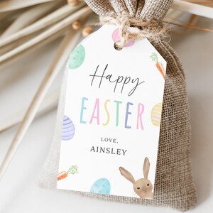 May include: A white gift tag with the text "Happy Easter" in colorful letters, decorated with pastel Easter eggs and carrots. The tag is tied to a brown burlap gift bag. The text "Love, Ainsley" is printed below the greeting.