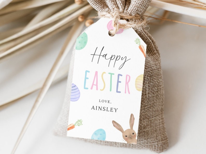 May include: A white gift tag with the text "Happy Easter" in colorful letters, decorated with pastel Easter eggs and carrots. The tag is tied to a brown burlap gift bag. The text "Love, Ainsley" is printed below the greeting.