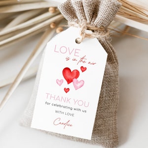 May include: A small burlap bag tied with twine, featuring a white tag with the text "Love is in the air" and "Thank you". The tag has watercolor hearts in shades of red and pink. A romantic gift or favor.