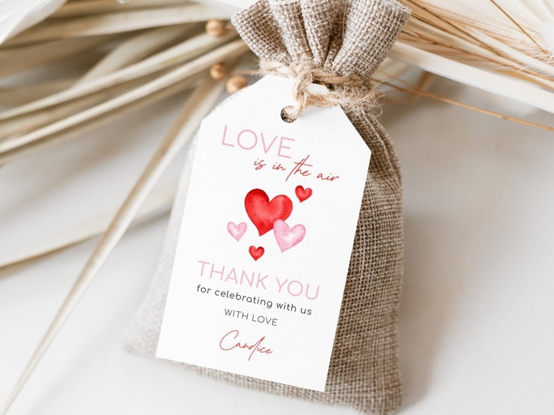 May include: A small burlap bag tied with twine, featuring a white tag with the text "Love is in the air" and "Thank you". The tag has watercolor hearts in shades of red and pink. A romantic gift or favor.
