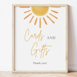 May include: A white sign with a yellow sun graphic and the text "Cards and Gifts" in gold script. The sign also includes the text "Thank you!" in black.