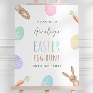 Puede incluir: Un cartel blanco con el texto "Bienvenido a la fiesta de cumpleaños de Ainsley's Easter Egg Hunt". El cartel está decorado con huevos de colores, zanahorias y conejos.