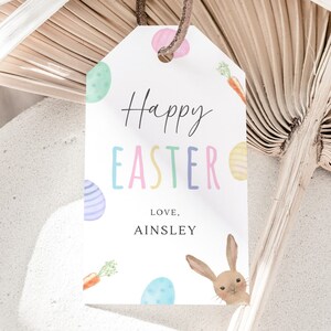 May include: White gift tag with the text "Happy Easter" in colorful letters. The tag is decorated with pastel colored eggs, carrots, and a bunny rabbit peeking out from the bottom right corner. The text "Love, Ainsley" is at the bottom of the tag.