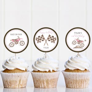 May include: Three cupcakes with white frosting, each topped with a circular decoration. The decorations feature a pink dirt bike, checkered flags, and the text "Race On Over", "Happy Birthday Tyler", and "Tyler's 3rd Birthday".