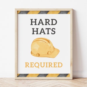 May include: A printable sign with black text on a white background that reads "Hard Hats Required". A yellow construction helmet is illustrated below the text. The sign is framed in a light wood frame with black and yellow striped tape around the edge.