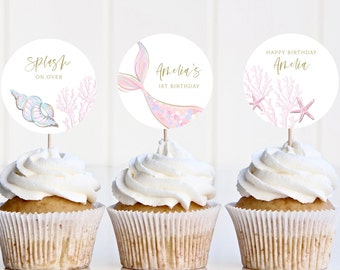 May include: Three cupcakes decorated with round, white paper toppers. The toppers feature a mermaid tail, a seashell and coral, and coral and starfish. The text on the toppers reads "Splash on Over", "Amelia's 1st Birthday", and "Happy Birthday Amelia".