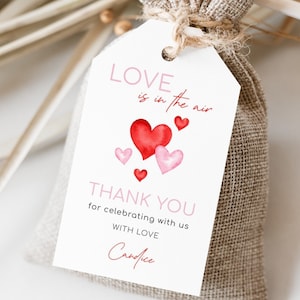 May include: A white gift tag with the text "LOVE is in the air" and "THANK YOU" in pink script, along with watercolor hearts in red and pink. The tag is tied to a burlap bag with twine.
