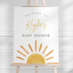 May include: Welcome to Kaylee's Baby Shower sign with a watercolor illustration of a yellow sun with orange rays on a white background.