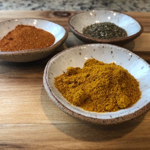 May include: Three small white bowls with rims, each filled with a different spice. The bowl on the left contains a red spice, the middle bowl contains a yellow spice, and the bowl on the right contains a green spice.