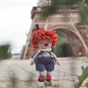 May include: A crocheted clown doll with red hair, a blue and white striped outfit, and a red nose. The doll is standing in front of a blurred background of a building.