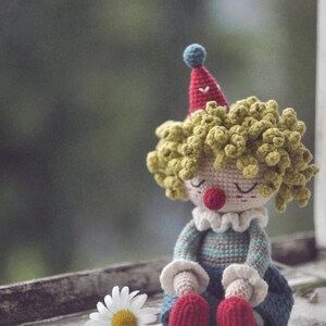 May include: A crocheted clown doll with yellow yarn hair, a red and blue hat, and a striped blue and gray outfit. The doll is sitting on a white surface with a white daisy.