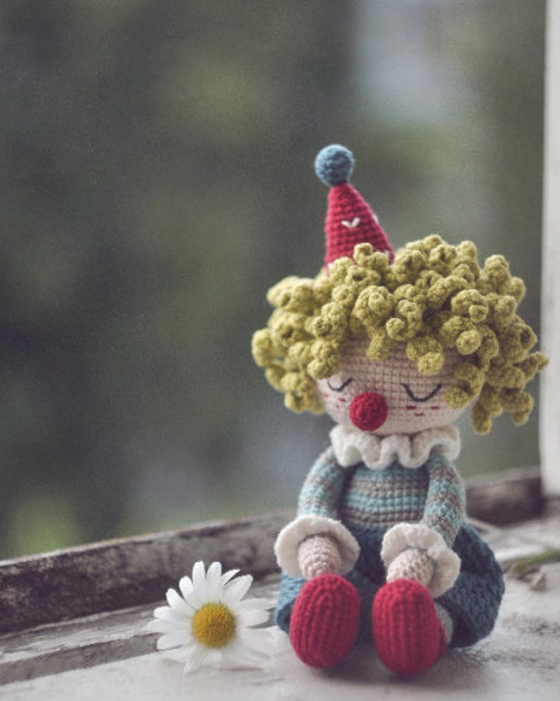 May include: A crocheted clown doll with yellow yarn hair, a red and blue hat, and a striped blue and gray outfit. The doll is sitting on a white surface with a white daisy.