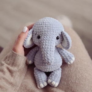 Puede incluir: Un juguete de elefante tejido de color azul claro con detalles blancos en las orejas y los pies. El animal de peluche hecho a mano tiene una superficie texturizada y pequeños ojos negros. Es un juguete suave.