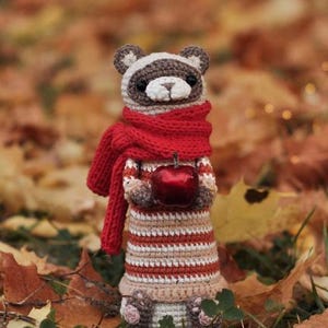 Puede incluir: Un mapache de crochet con una bufanda roja y sosteniendo una manzana roja. El mapache est&aacute; de pie sobre una cama de hojas de oto&ntilde;o.
