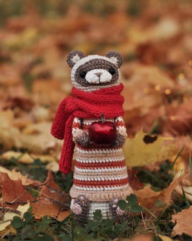 Puede incluir: Un mapache de crochet con una bufanda roja y sosteniendo una manzana roja. El mapache est&aacute; de pie sobre una cama de hojas de oto&ntilde;o.