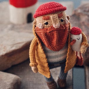 Könnte beinhalten: Eine gehäkelte Puppe einer bärtigen Person mit roter Mütze, gelber Jacke und gestreiftem Oberteil. Die Puppe hat einen großen orangefarbenen Bart, rosa Wangen und hält einen kleinen weißen Vogel mit roter Kappe und orangefarbenem Schnabel.