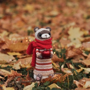 Puede incluir: Un animal de peluche tejido a crochet que se asemeja a un hur&oacute;n con una bufanda roja y sosteniendo una manzana roja. El animal est&aacute; de pie sobre una cama de hojas de oto&ntilde;o.