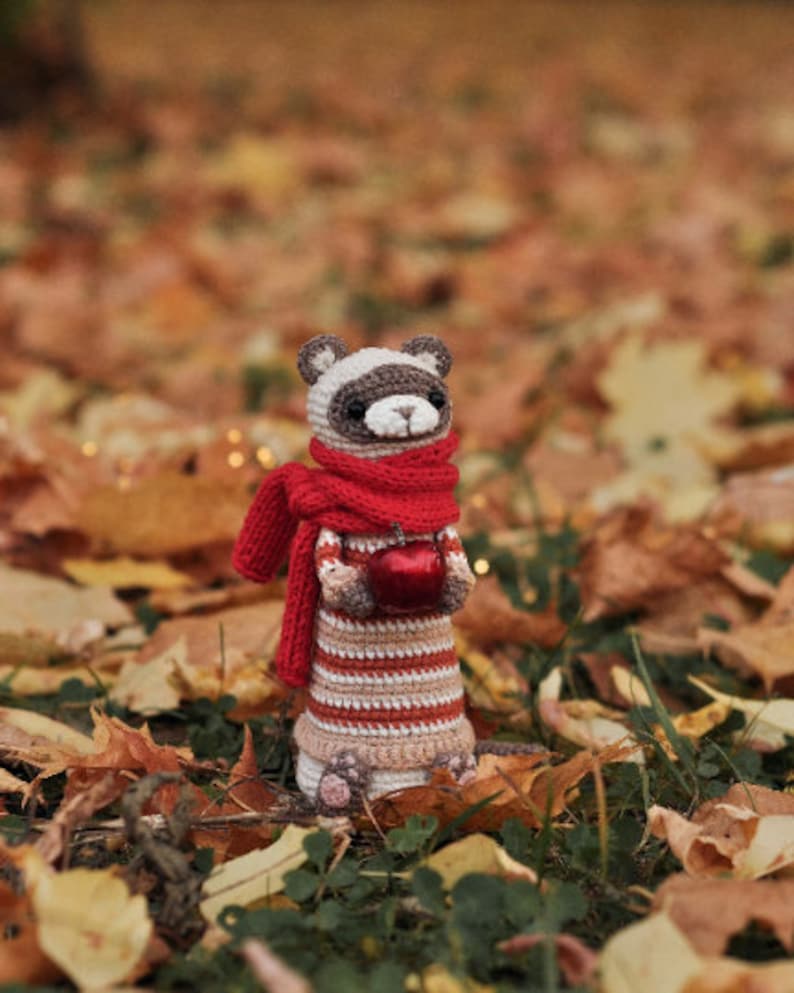 Puede incluir: Un animal de peluche tejido a crochet que se asemeja a un hur&oacute;n con una bufanda roja y sosteniendo una manzana roja. El animal est&aacute; de pie sobre una cama de hojas de oto&ntilde;o.