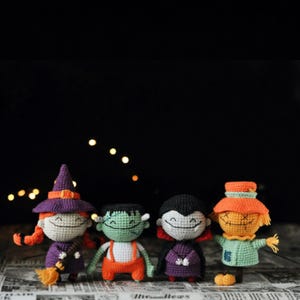 May include: Four small crocheted dolls featuring Halloween characters. From left to right: a witch with a purple hat and broom, a green monster with orange overalls, a vampire in a black cape, and a scarecrow with an orange hat and light green shirt.