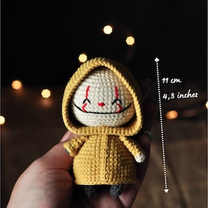 Könnte beinhalten: Eine gehäkelte gelbe Figur in einem Regenmantel mit weißem Gesicht und rotem Lächeln. Die Figur ist 11 cm groß.
