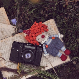 May include: A crocheted clown doll with red hair and a blue and white striped shirt lies on an open book next to a black Zenit camera.