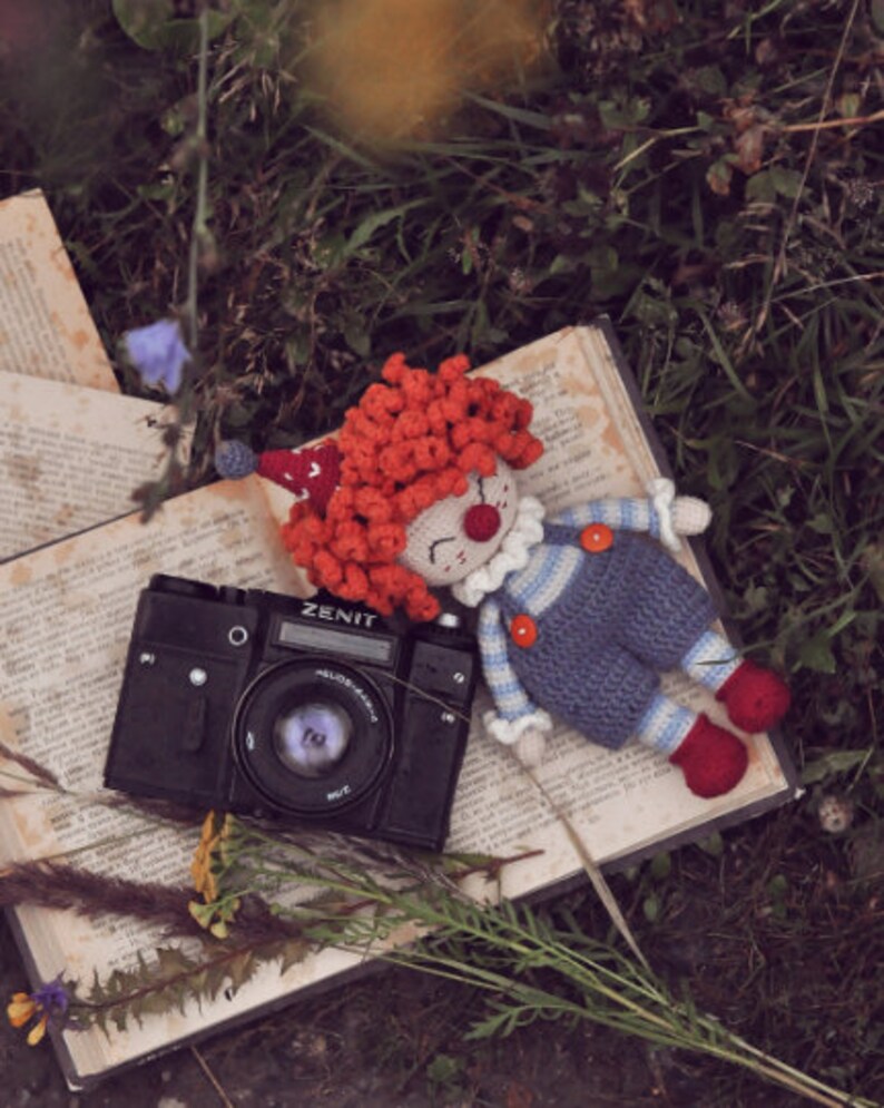 May include: A crocheted clown doll with red hair and a blue and white striped shirt lies on an open book next to a black Zenit camera.