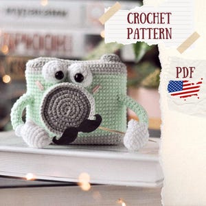 May include: Crochet pattern PDF for a green and gray camera amigurumi toy with a black mustache. The pattern includes instructions for making the camera, eyes, and mustache.