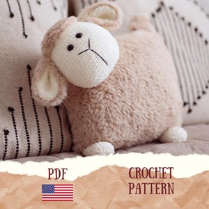 Op de afbeelding: Een zacht, lichtbruin schaapvormig kussen met een wit gehaakt gezicht, oren en voeten. Het schaap heeft zwarte ogen en een zwart gestikte mond. De afbeelding bevat ook de tekst "PDF" en "CROCHET PATTERN" met de Amerikaanse vlag.