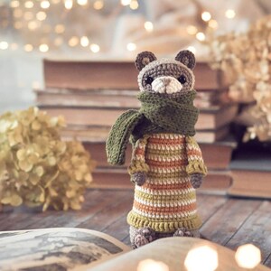 Puede incluir: Un animal de peluche tejido a crochet con una bufanda verde y un vestido a rayas. El animal est&aacute; sentado en un libro con un fondo borroso de luces y libros.