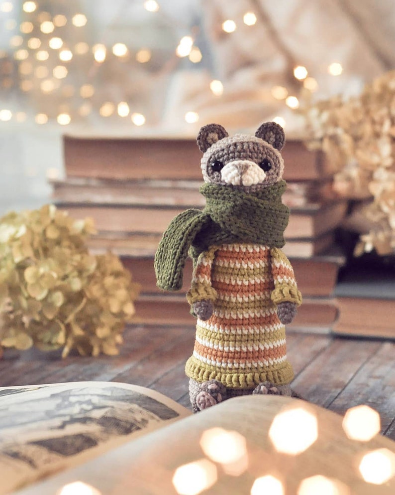 Puede incluir: Un animal de peluche tejido a crochet con una bufanda verde y un vestido a rayas. El animal est&aacute; sentado en un libro con un fondo borroso de luces y libros.