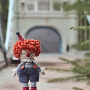 May include: A crocheted clown doll with red hair, a blue and white striped outfit, and a red nose. The doll is standing on a gray surface.